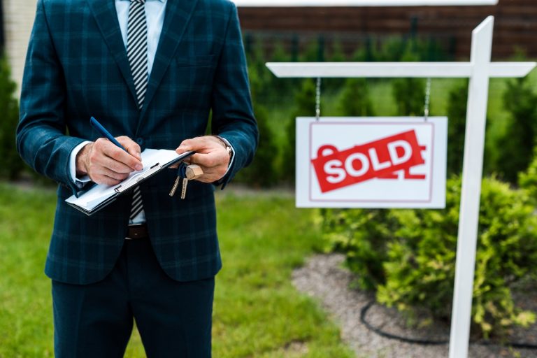 A real estate agent standing with a clipboard next to a sign with a "SOLD" sticker across it.